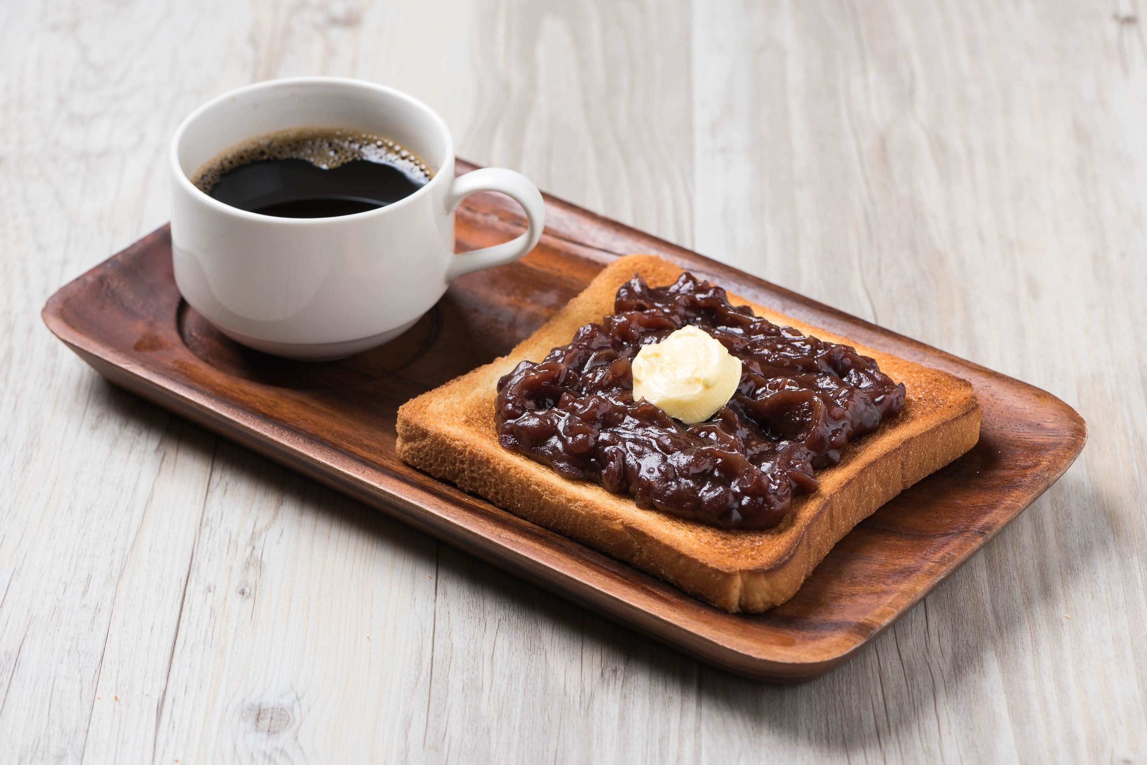 A slice of toast with jam and a pat of butter on a wooden tray, accompanied by a cup of coffee.