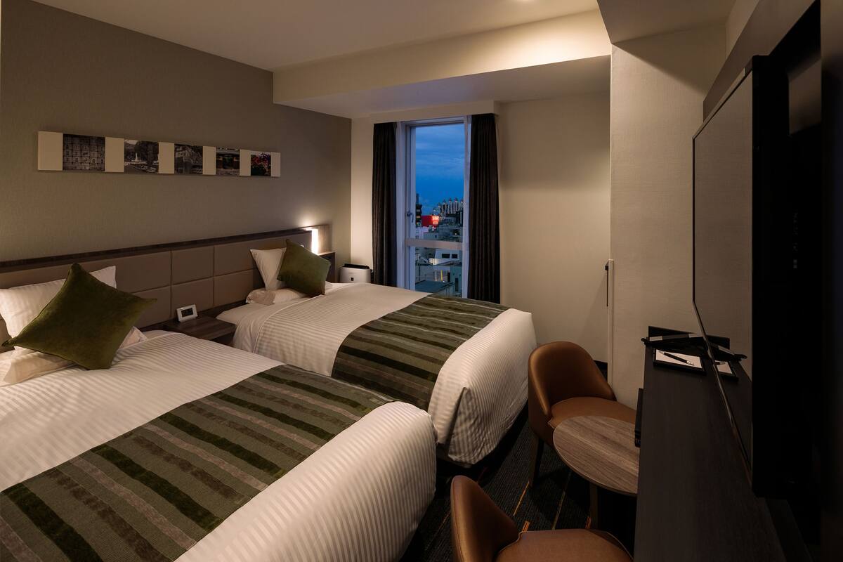 A hotel room with two beds, a TV, a desk, and a view of the city.