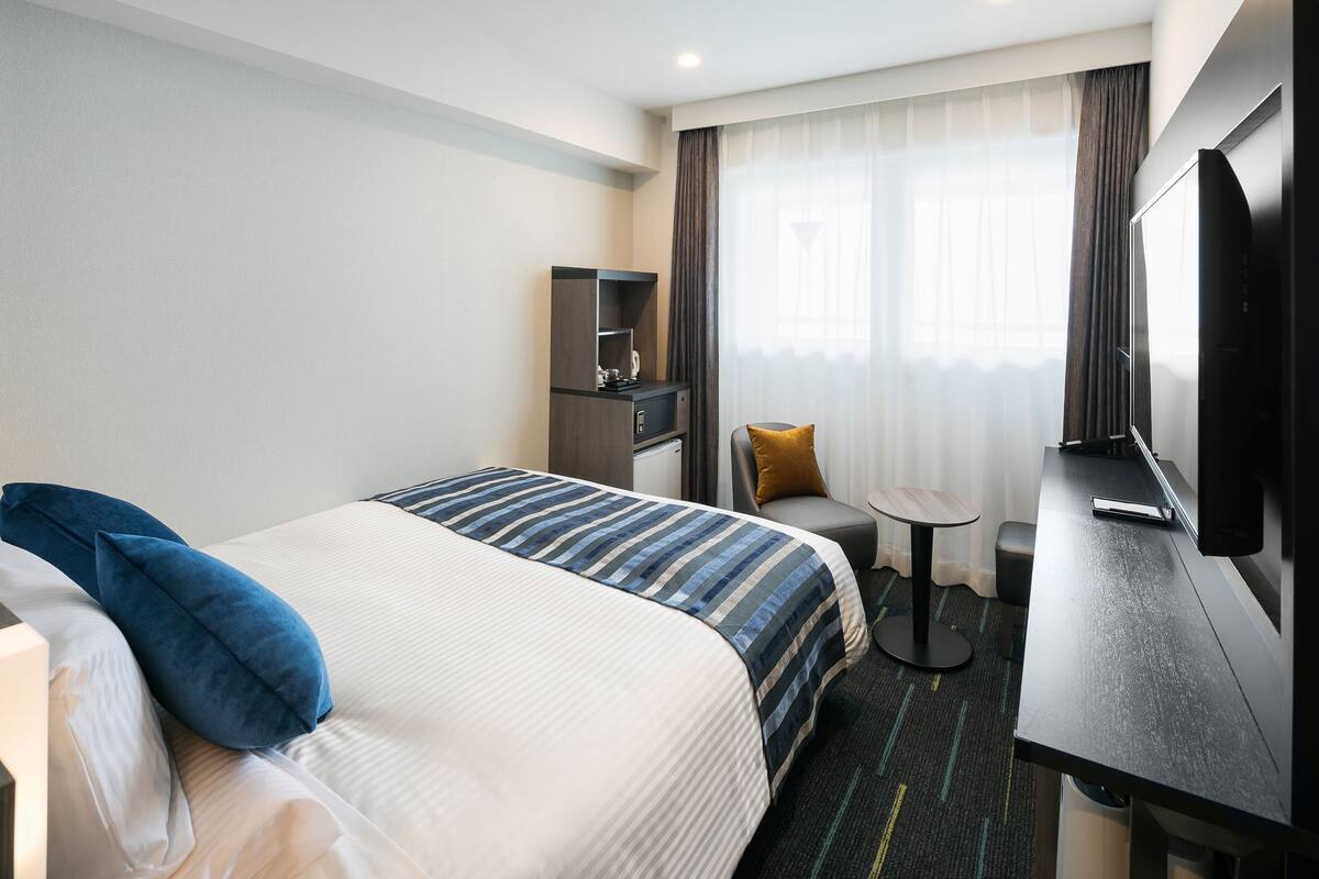 A hotel room with a bed, a desk, a chair, a TV, and a small table.