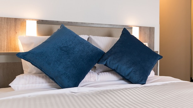 Ein ordentlich bezogenes Bett mit weißer Bettwäsche und zwei blauen Kissen, ein hölzerner Kopfteil und eine wandmontierte Leuchte.