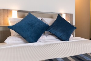 A neatly made bed with white bedding and two blue pillows, a wooden headboard, and a wall-mounted light fixture. - Hotel Mystays Nagoya Nishiki (Nagoya)