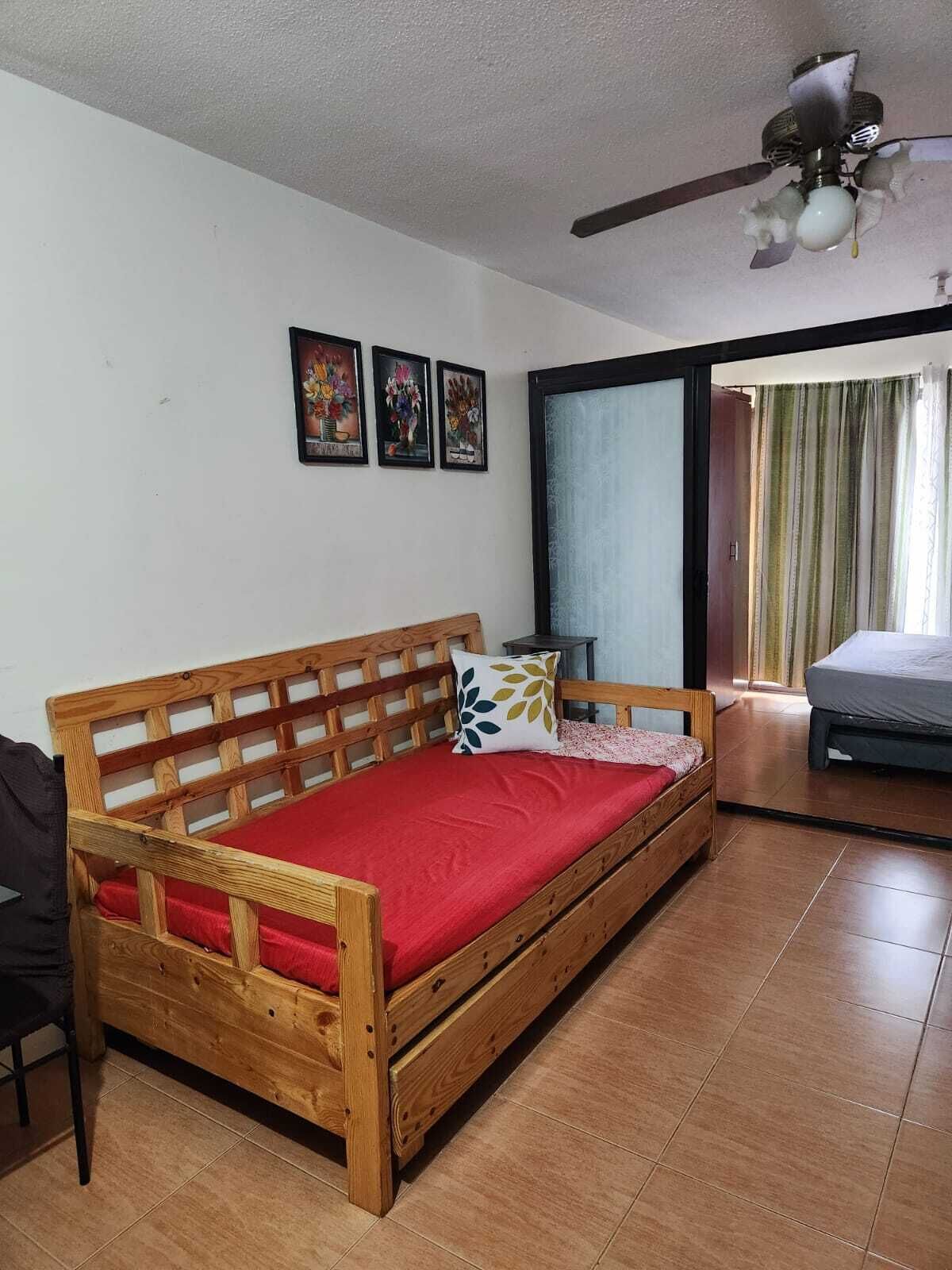 Una cama de madera con colchón rojo y cabecera de madera en una habitación con suelo de baldosas, ventilador de techo y tres cuadros en la pared.