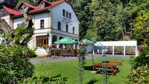 A hotel with a white facade, red roof, and a sign indicating it is a hotel. There are outdoor tables and umbrellas, suggesting a dining or relaxation area. - Hotel Pod Šikmou Věží (Holín)