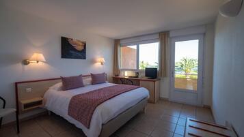 A bedroom with a bed, bedside tables, a desk, a chair, a TV, and a window with a view of palm trees.