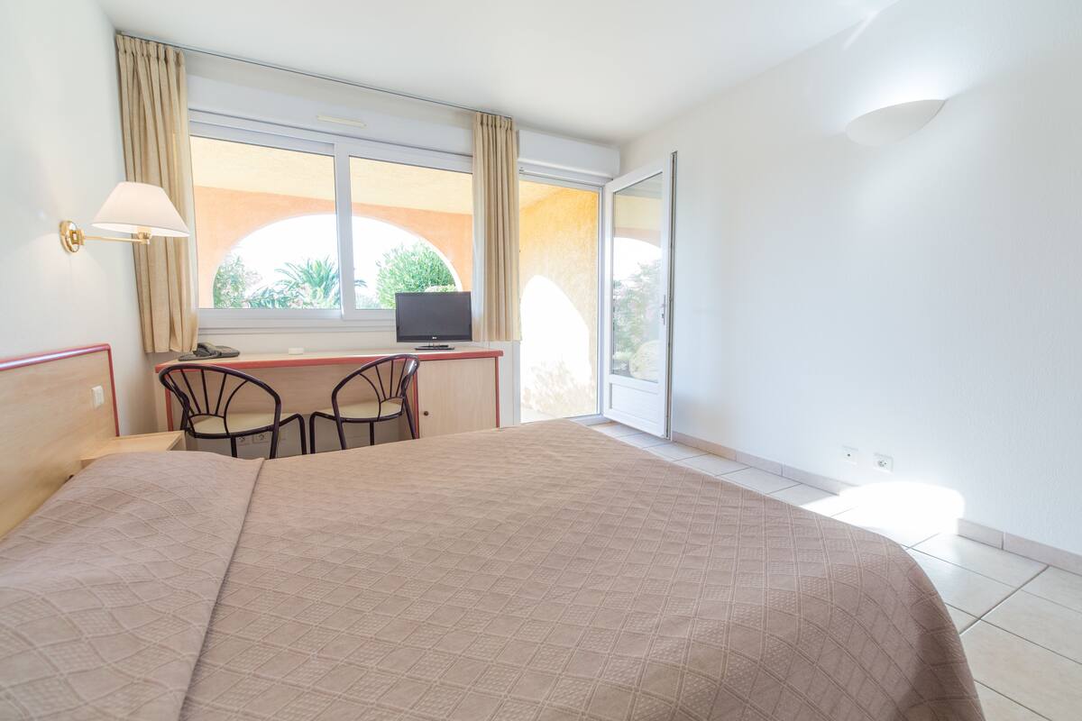 A hotel room with a bed, a desk with a computer, two chairs, and a window with curtains.