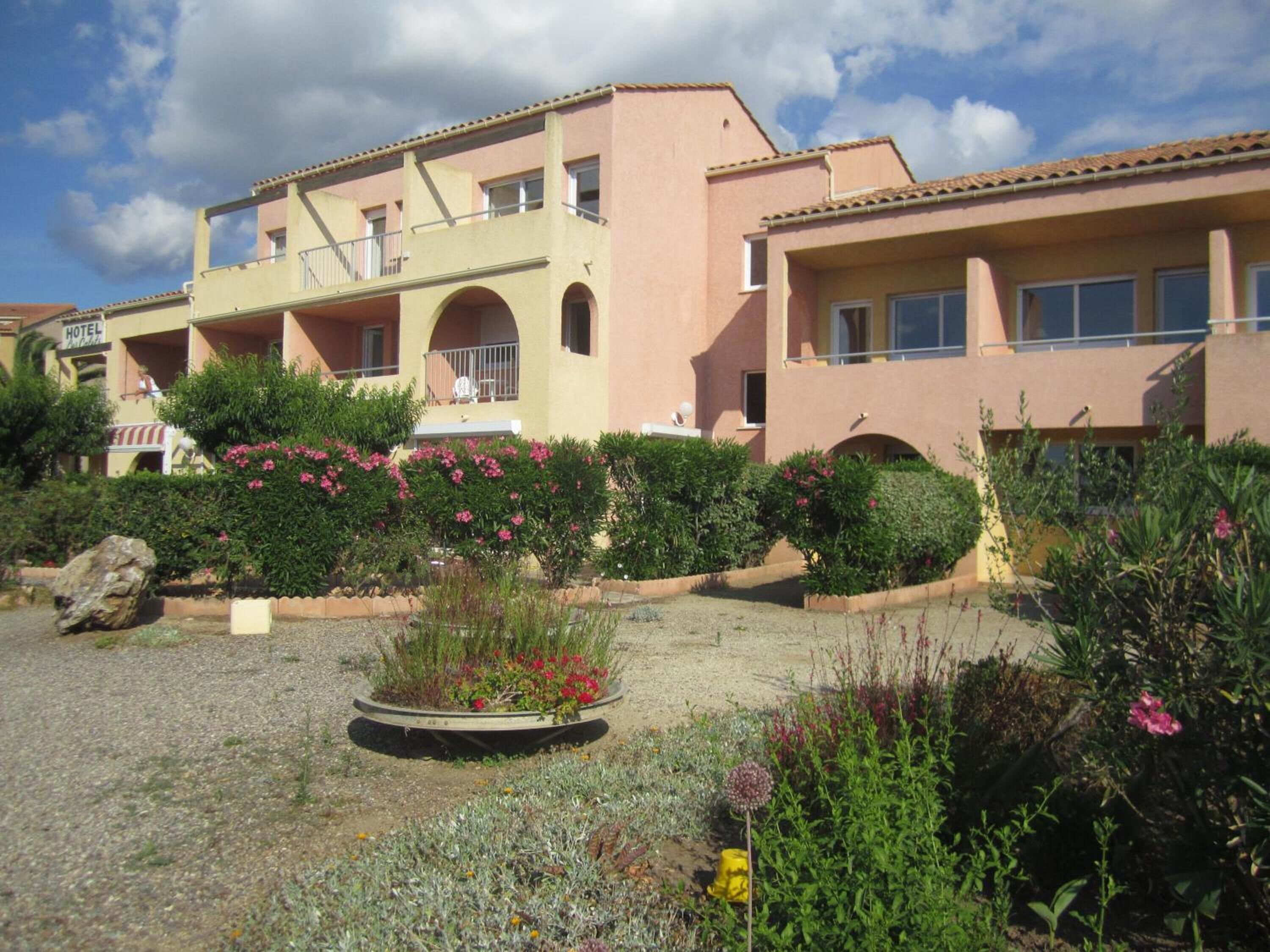 Ein Hotel mit rosa Fassade, mehreren Balkonen und einem Gartenbereich mit verschiedenen Pflanzen und Blumen.