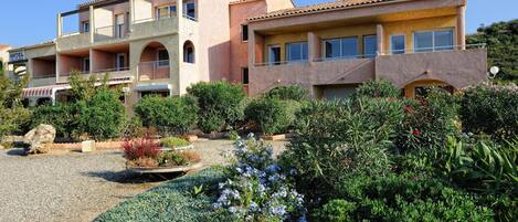 A hotel with multiple balconies, a garden with various plants and flowers, and a clear blue sky.