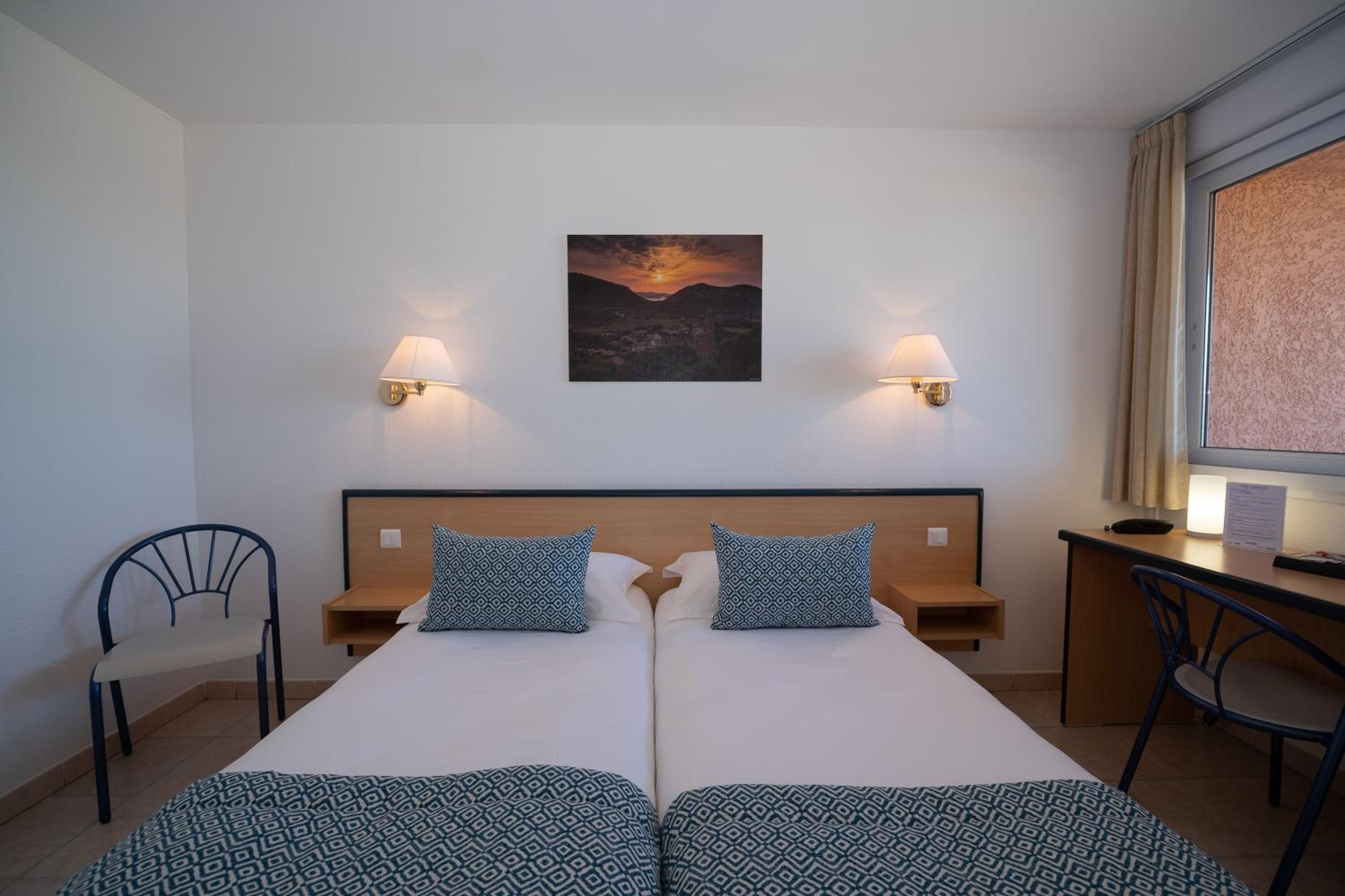 A double room with two beds, a desk, a chair, and a wall-mounted painting.