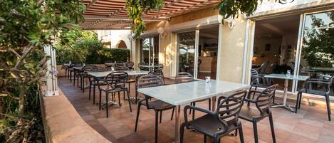 An outdoor patio with multiple tables and chairs, surrounded by greenery and a pergola.