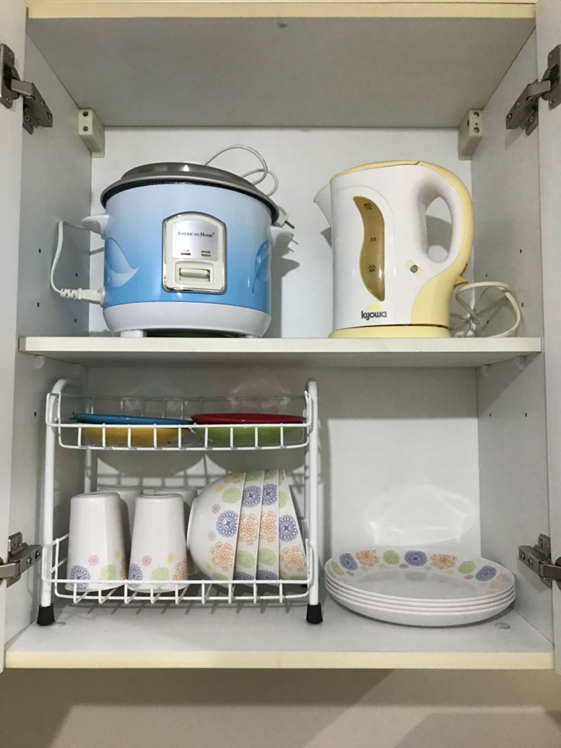Een keukenkastje met een blauwe slowcooker, een gele elektrische waterkoker, een afdruiprek met kopjes en borden, en een wit plankje.