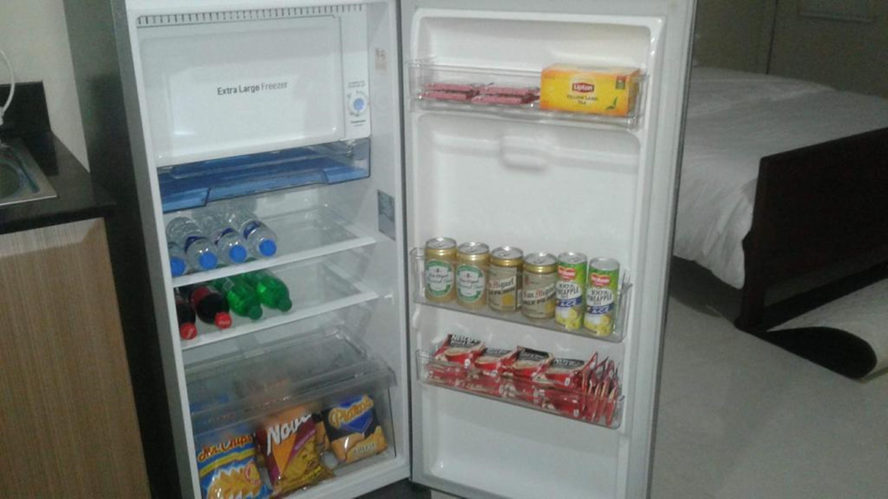 Ett hotellrum med ett kylskåp som står öppet och innehåller drycker och snacks, och en säng syns i bakgrunden.