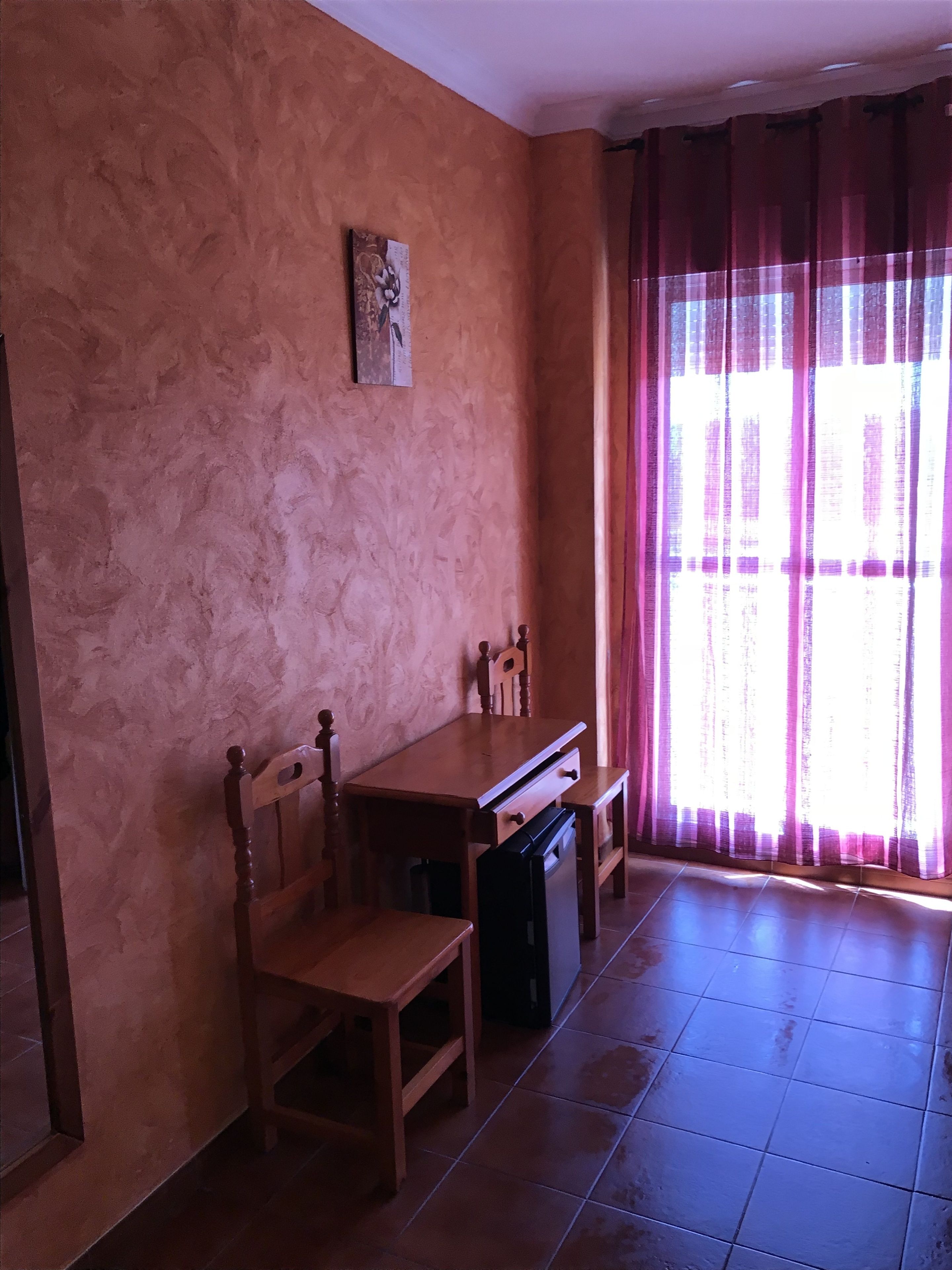 A room with a wooden dining table and chairs, a small painting on the wall, and a window with pink curtains.