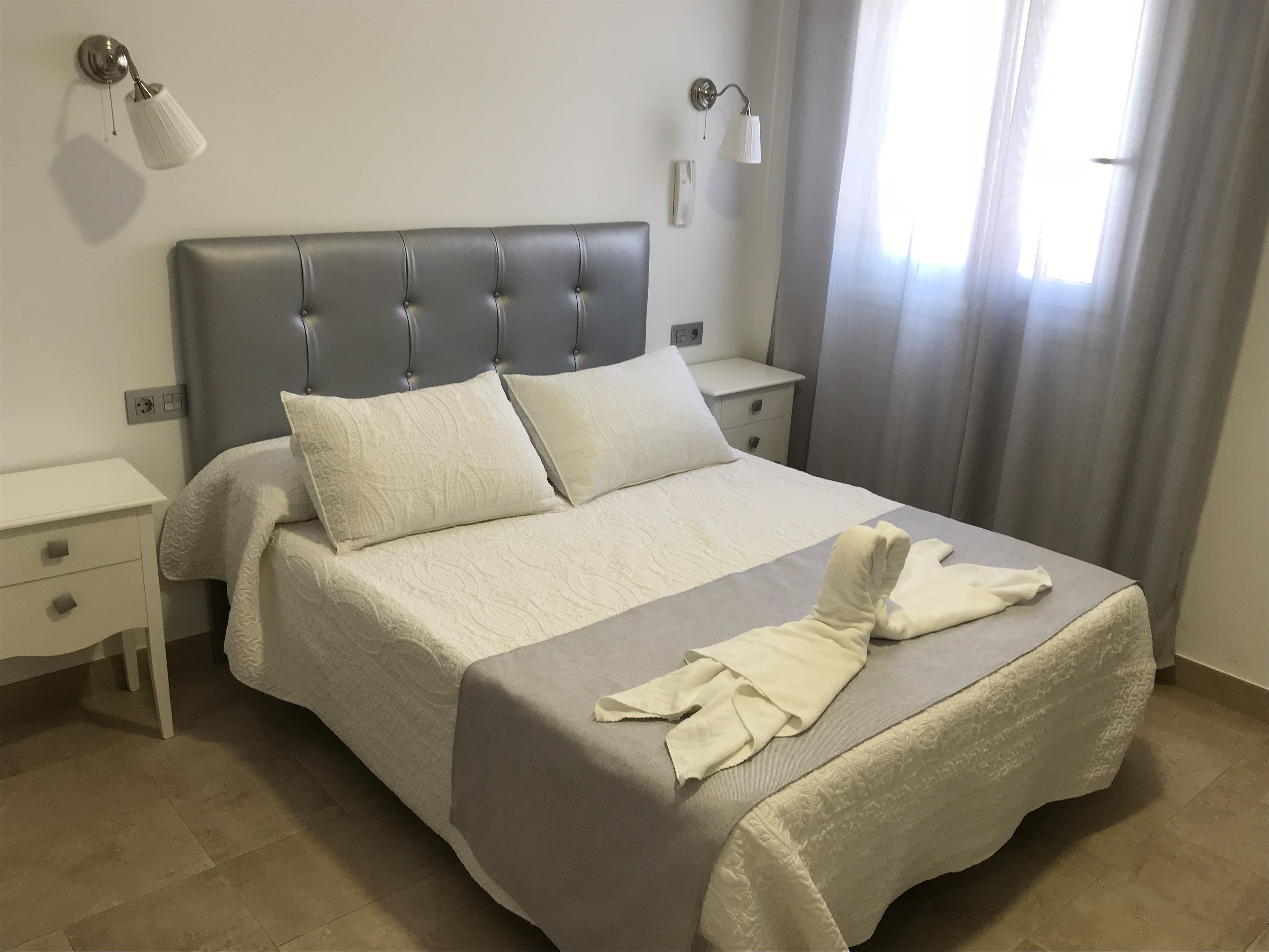 A neatly made bed with a grey headboard, white bedding, and a folded towel on the footboard. There are two bedside tables with lamps, a nightstand, and a window with curtains.