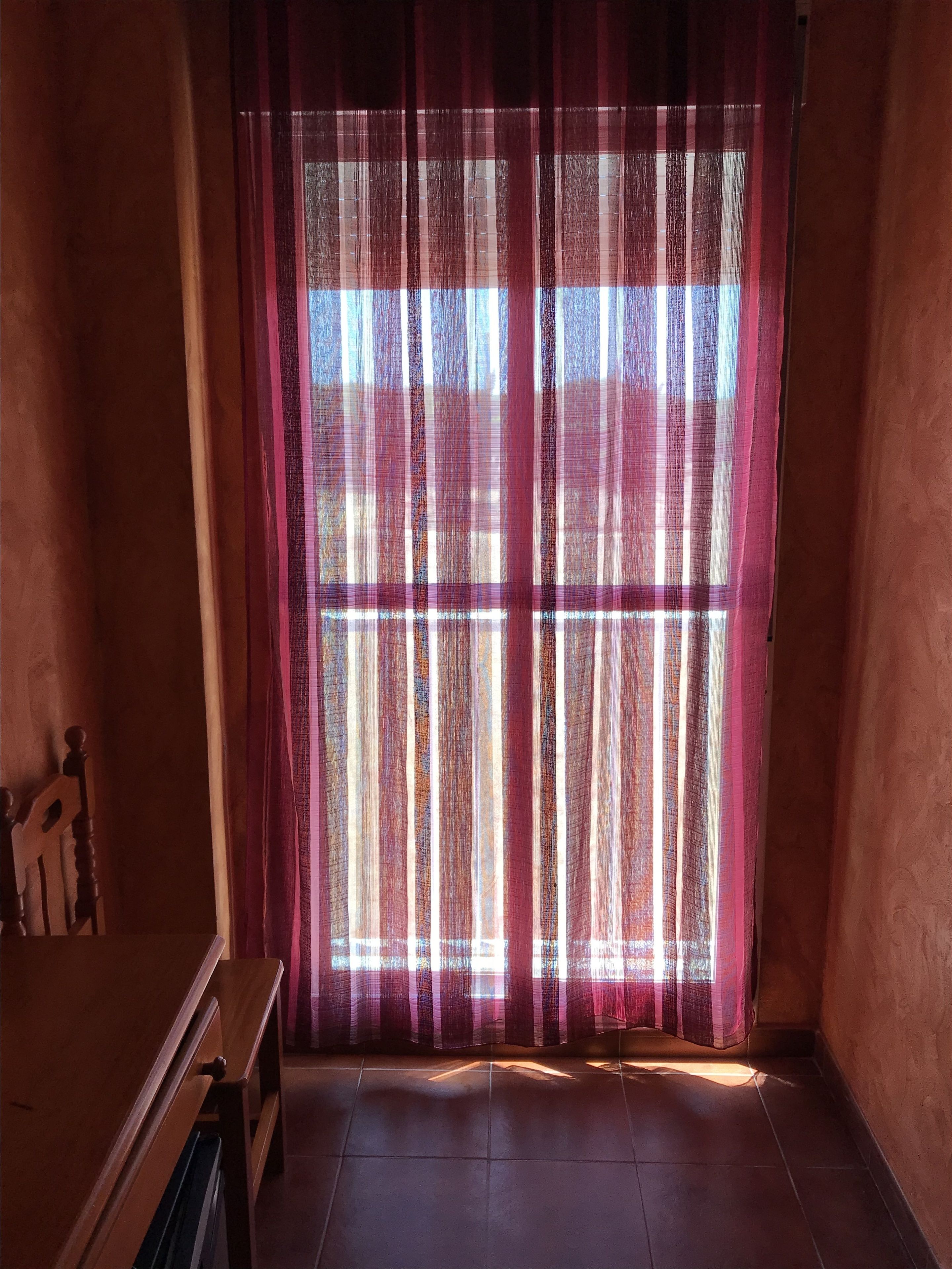 Una habitación con un ventanal grande cubierto por una cortina translúcida, muebles de madera y piso de baldosas.