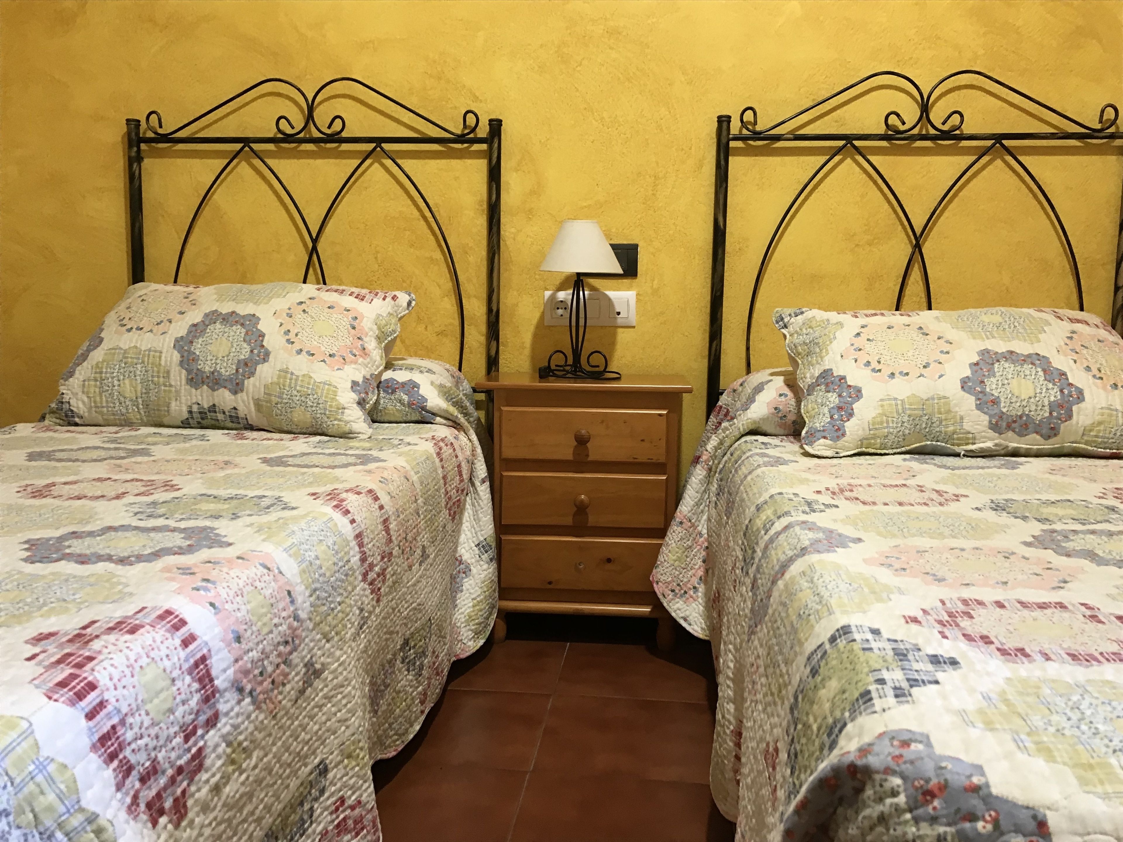Una habitación con dos camas, cada una con un edredón estampado y almohadas a juego, una mesita de noche de madera con lámpara y suelo de baldosas.