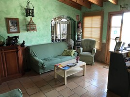 Una sala de estar con un sofá verde, una mesa de centro de madera con libros, un sillón verde, un ventilador y un extintor.