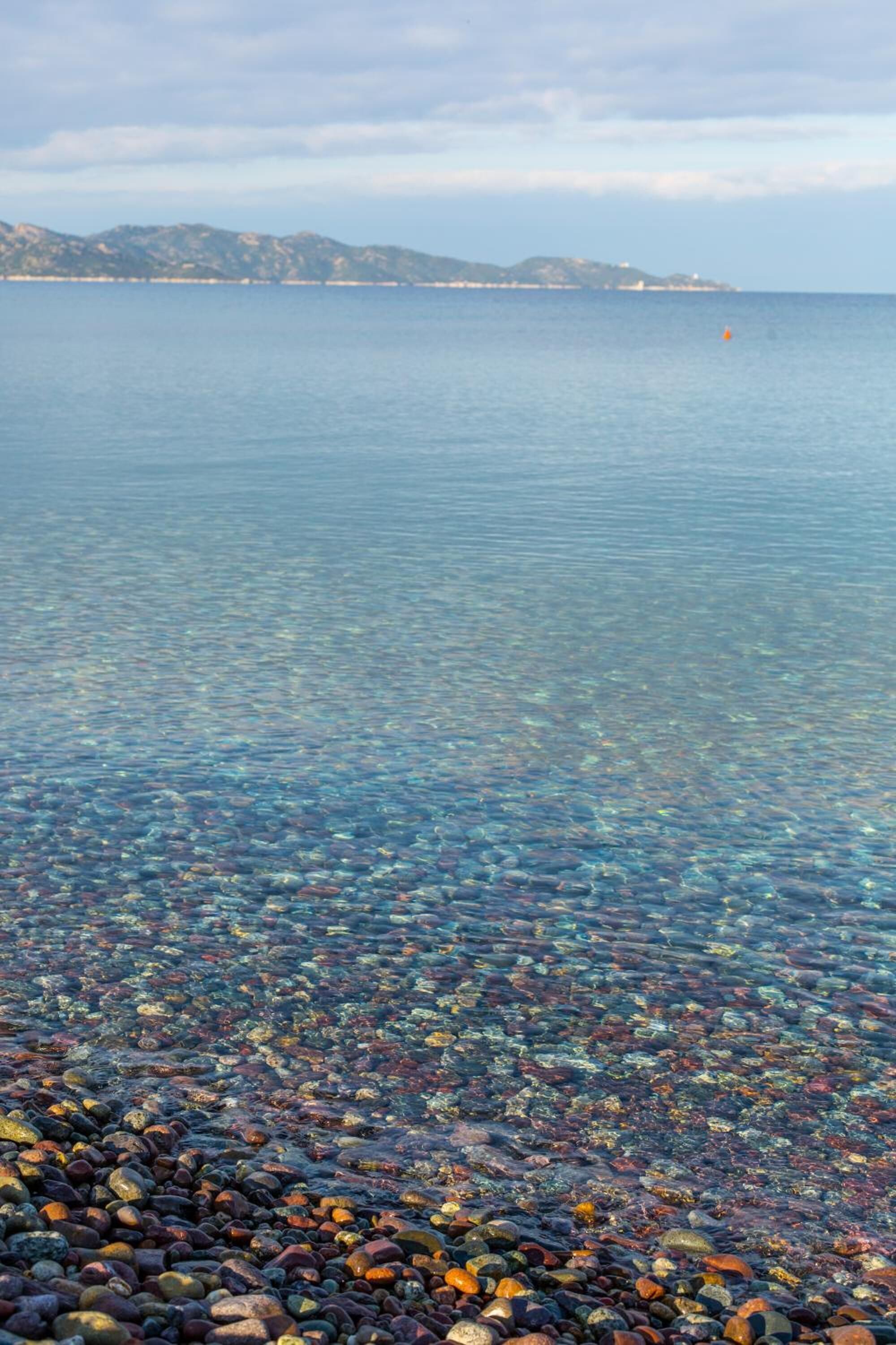 A clear view of a pebble beach with a calm sea and distant landforms.