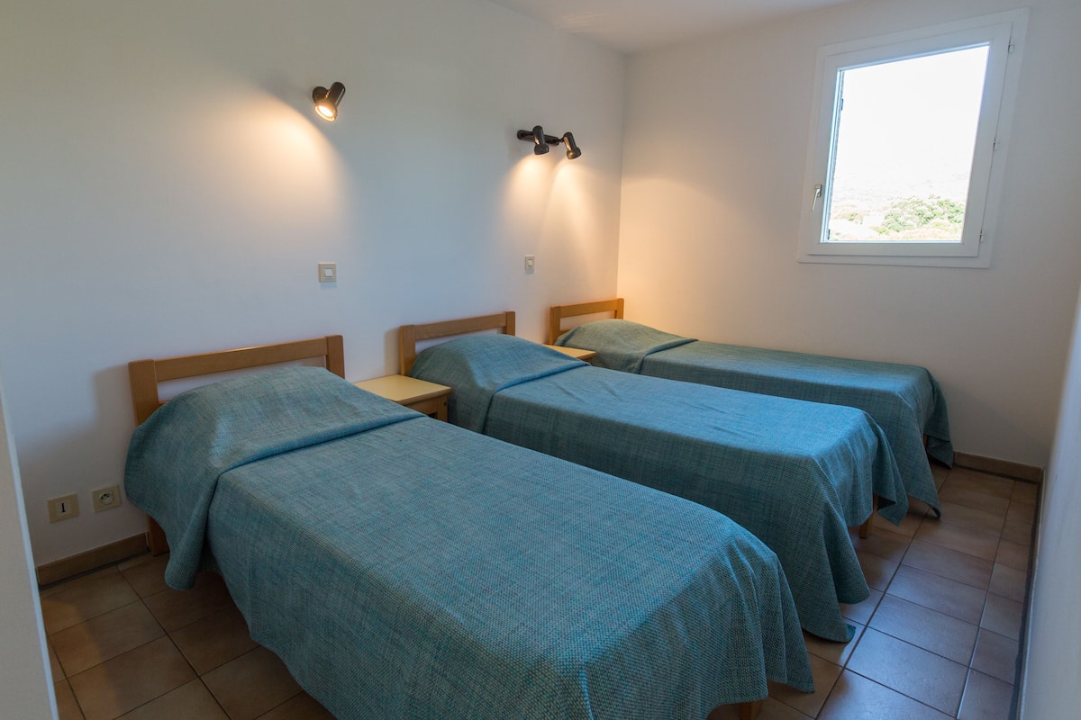 A room with three single beds, each with a wooden headboard and a teal bedspread. There are wall-mounted lights above each bed and a window on the right wall.
