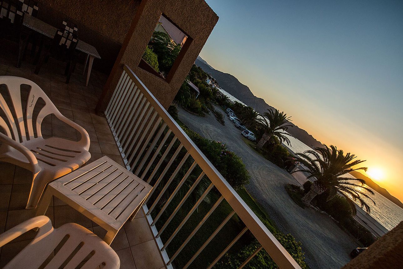 A balcony with chairs and a table, overlooking a coastal road with parked cars and palm trees, and a mountain in the background.