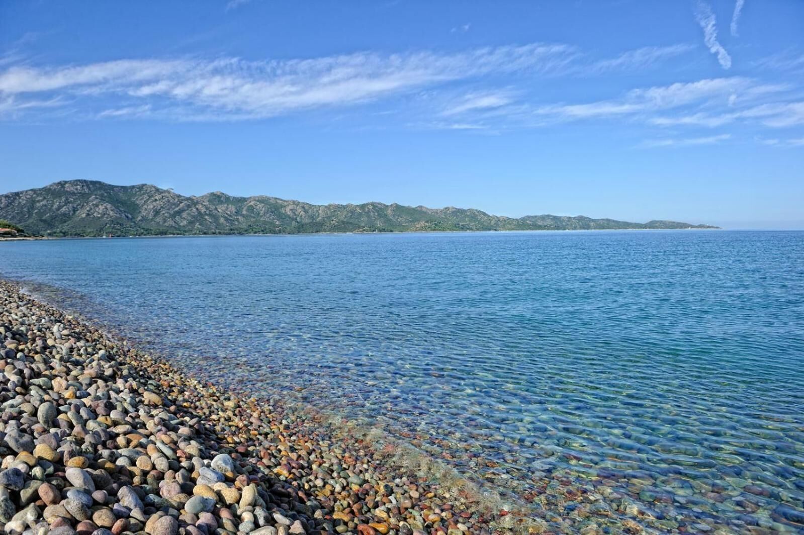 A pebbled beach with clear water and distant mountains under a blue sky.
