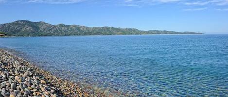 A pebbled beach with clear water and distant mountains under a blue sky.
