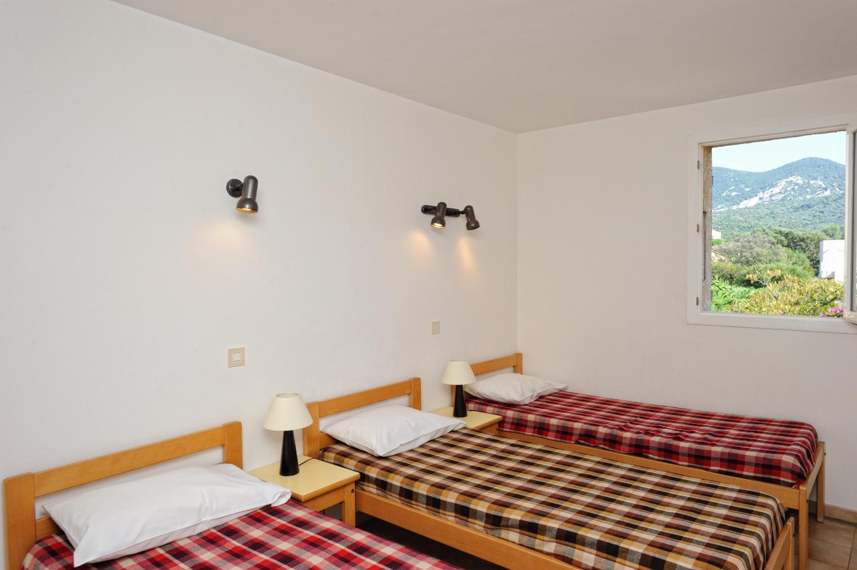 Una habitación con tres camas de madera, cada una con colcha a cuadros blancos y rojos y almohadas blancas. Hay mesitas de noche con lámparas, y las paredes son de color blanco. Una ventana al lado derecho ofrece vistas a la vegetación y montañas.