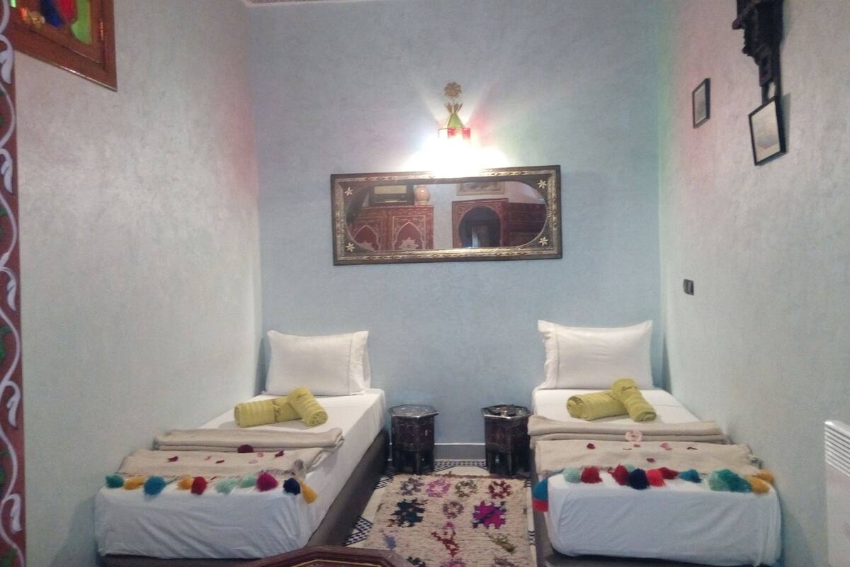 A small hotel room with two single beds, a mirror, a rug, and a wall-mounted light fixture.
