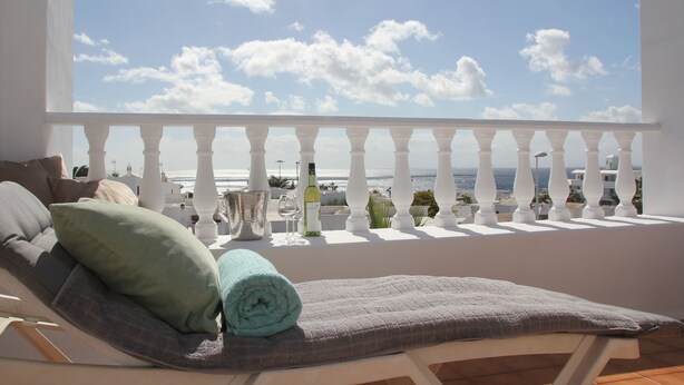 A balcony with a view of the sea, a lounge chair with cushions, and a bottle of wine. - Spacious two bedroom house near the beach with private terrace and sea views (Puerto del Carmen)