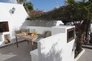 Une terrasse extérieure avec une table en bois, des coussins et un petit coin cuisine.