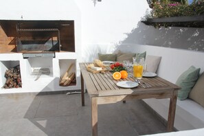 Une terrasse extérieure avec un barbecue, une table en bois, de l’assiette, des couverts et un verre de jus d’orange.