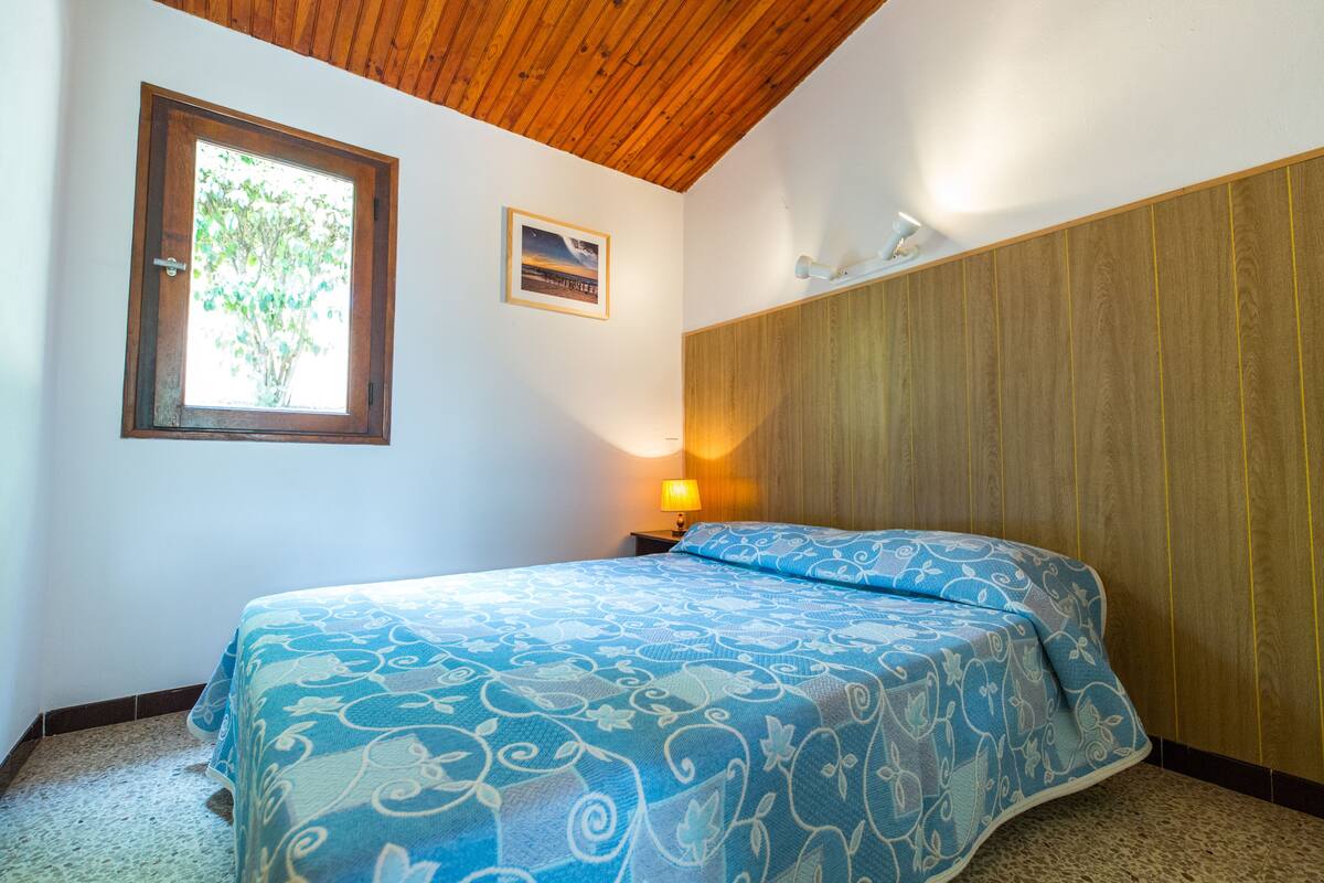Une chambre avec un plafond en bois, un lit recouvert d’une couvre-lit à motifs bleus, une fenêtre donnant sur la verdure et une lampe fixée au mur.