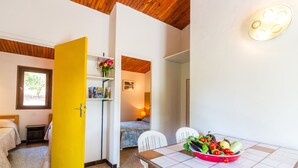 A compact room with a bed, a small table with a fruit bowl, a yellow door, and a window with a view of greenery. - Residence Les Arbousiers (Oletta)