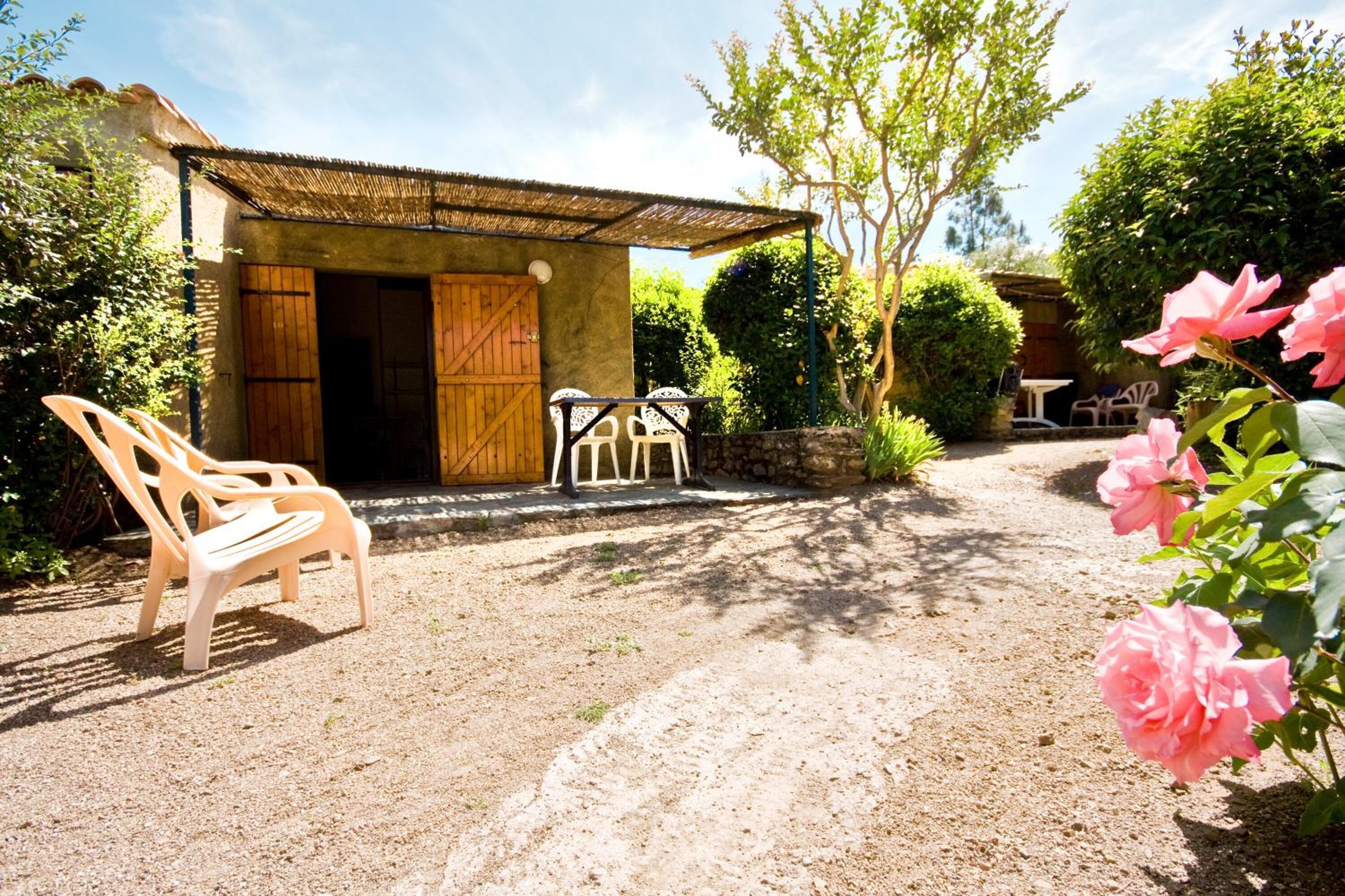 Un bâtiment rustique avec une porte en bois et un toit de chaume, entouré de verdure et bordé d’un chemin de gravier.