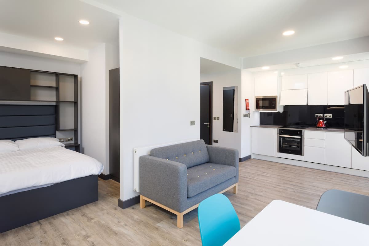 A modern hotel room with a bed, a grey sofa, a small table, and a kitchenette.