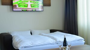 Une chambre d’hôtel avec un lit, un téléviseur fixé au mur qui diffuse une scène de jardin, et une table avec une bouteille et des verres.