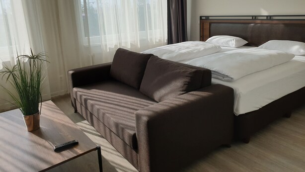 A hotel room with a bed, a brown sofa, a wooden coffee table, and a potted plant. - Motel Plus Schönefeld (Schoenefeld)