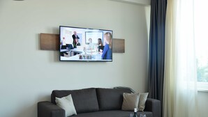 A modern living room with a large flat-screen TV mounted on the wall, a dark grey sofa with cushions, a wooden coffee table with a bottle of wine, a glass, and a remote control. - Motel Plus Schönefeld (Schoenefeld)
