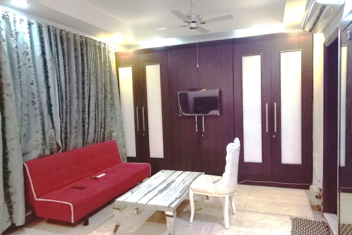 Una habitación con un sofá rojo, una mesa de centro blanca, un televisor montado en un mueble de madera oscura y un ventilador de techo.