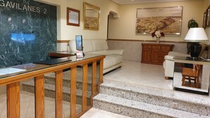 A hotel lobby with a marble reception desk, a sofa, a coffee table, a wooden cabinet, and a staircase with granite steps. - Hostal Los Gavilanes 2 (Toledo)