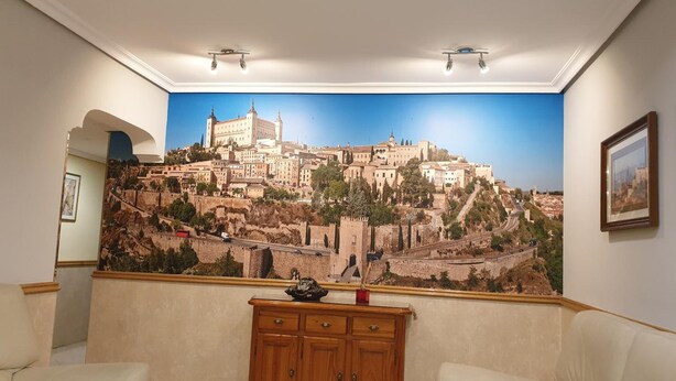A living room with a large cityscape mural, a wooden cabinet, and two sofas. - Hostal Los Gavilanes 2 (Toledo)