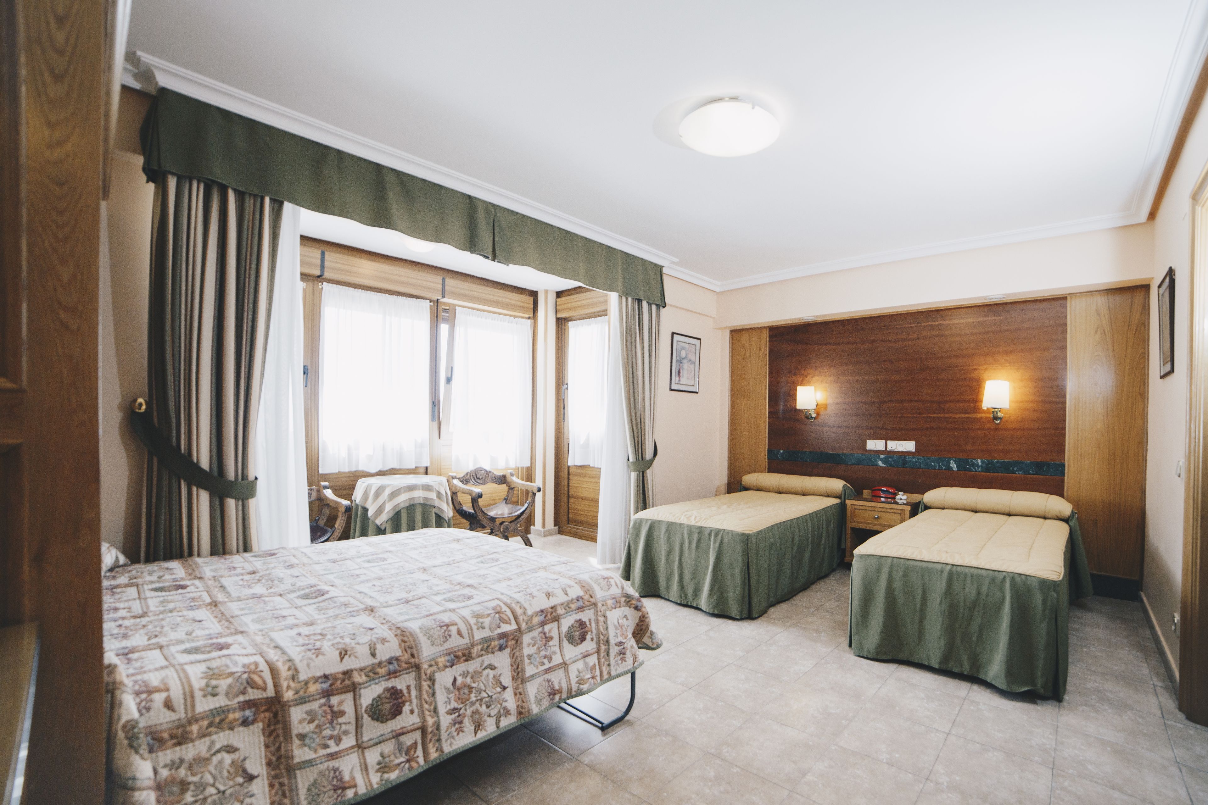 A hotel room with two single beds, a large bed, a wooden headboard, a window with curtains, and a wall-mounted lamp.