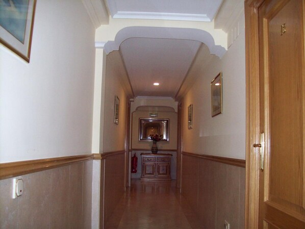 A hallway with a wooden door on the right, a fire extinguisher on the left, and a mirror on the wall. - Hostal Los Gavilanes 2 (Toledo)