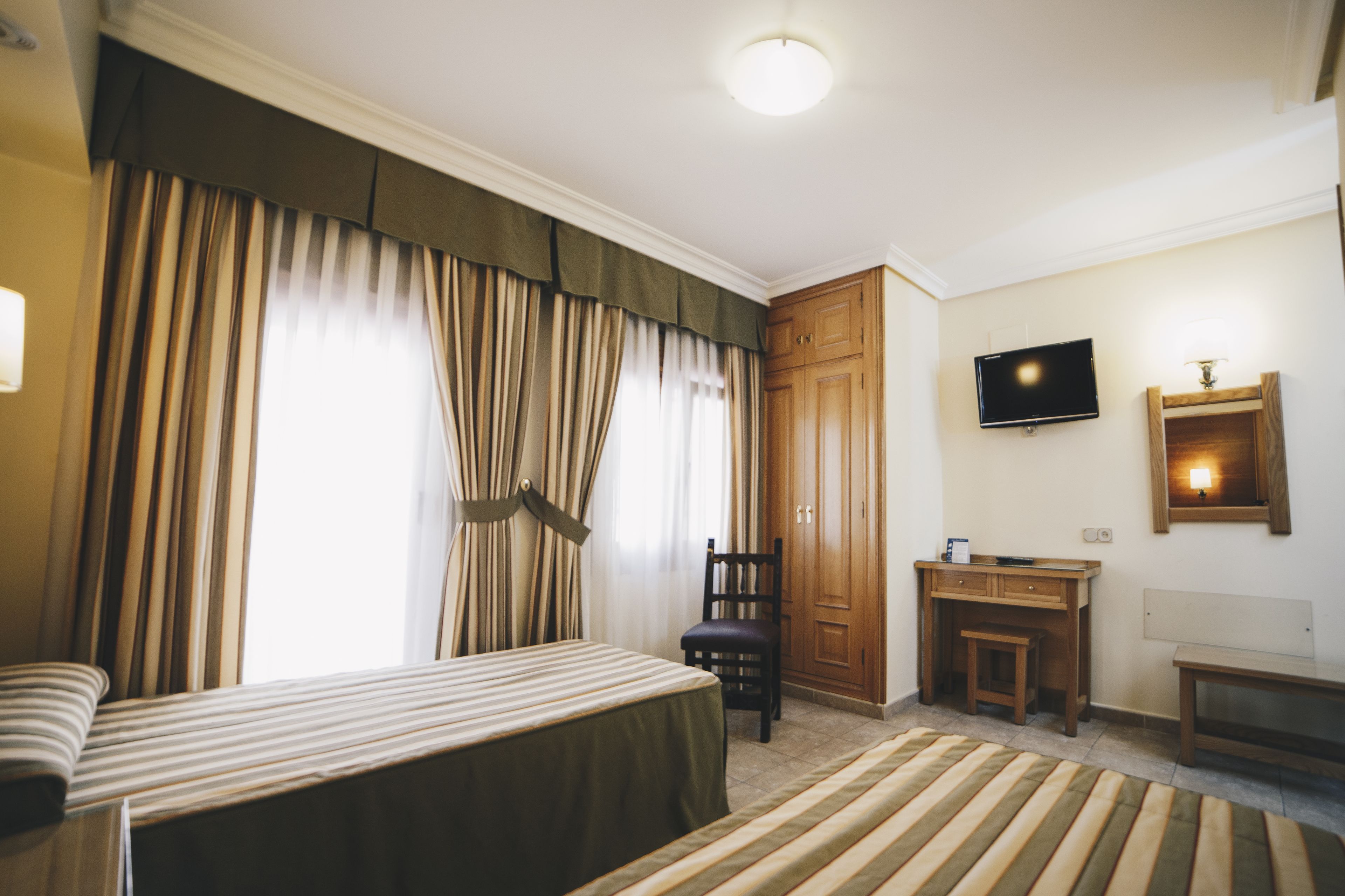 Quarto de hotel com cama, escrivaninha, cadeira, televisão e cortinas.