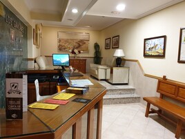 Un vestíbulo de hotel con mostrador de recepción, monitor de computadora, sofá, mesa de centro y una escalera que sube a otro piso.