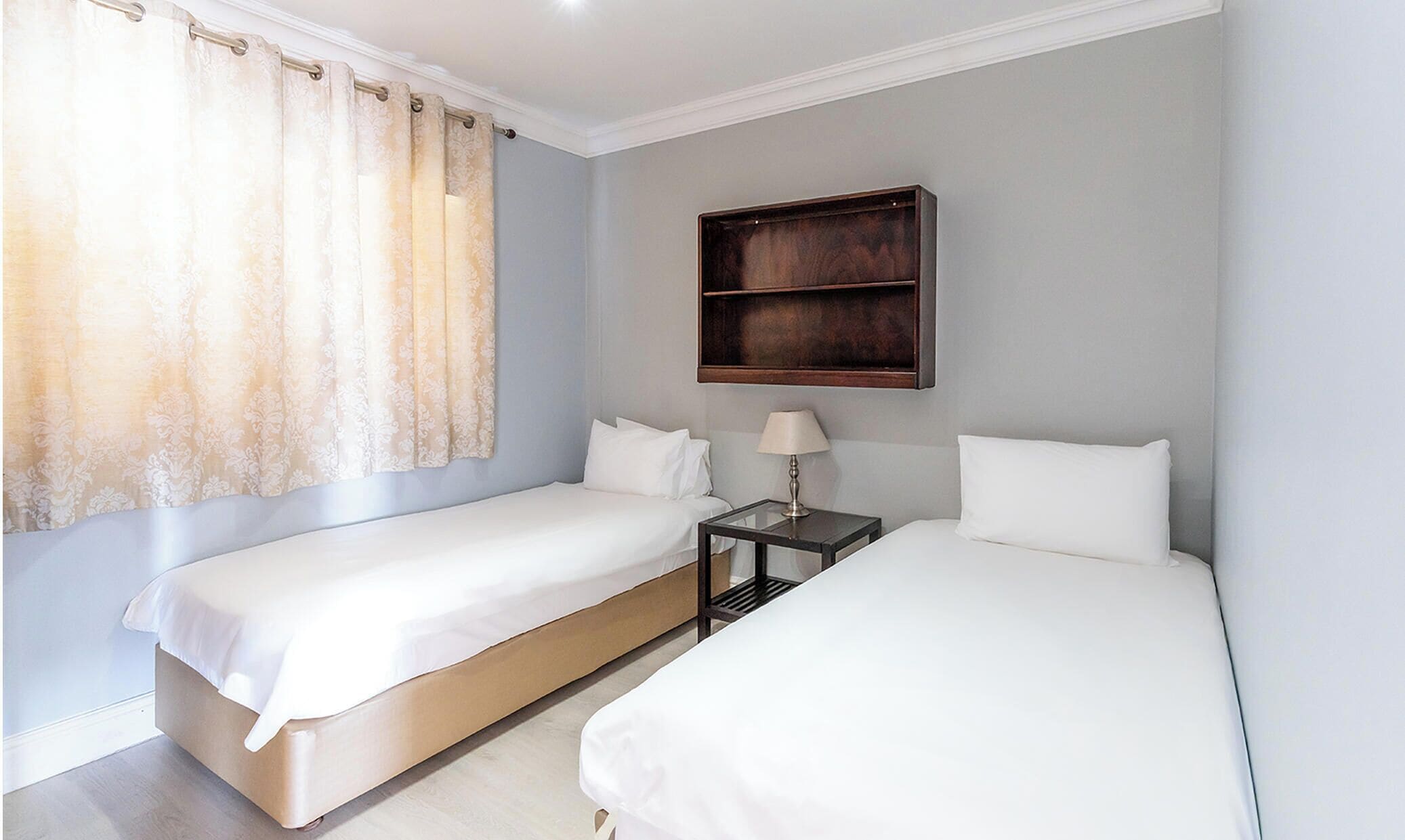 Quarto de hotel com duas camas de solteiro, uma mesinha de cabeceira com luminária e uma estante de madeira sobre as camas.