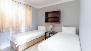 Quarto de hotel com duas camas de solteiro, uma mesinha de cabeceira com luminária e uma estante de madeira sobre as camas.