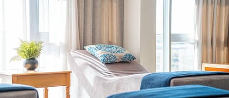 A hotel room with a bed, a bedside table with a plant, and a view of the city through large windows.