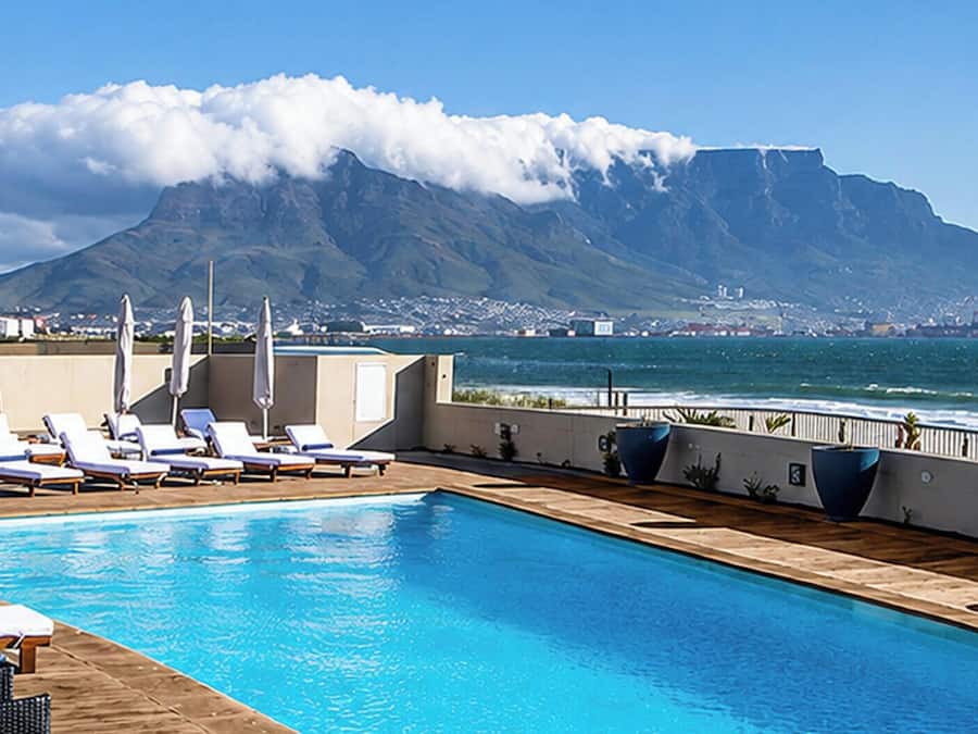 Une piscine bordée de chaises longues et dotée de parasols, offrant une vue sur un plan d’eau et une montagne.