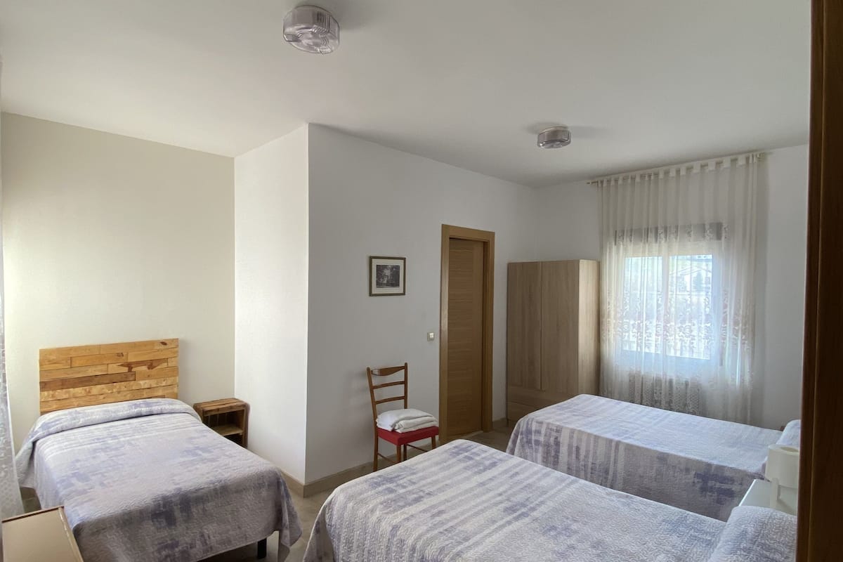 A room with two beds, a wooden headboard, a small table, a chair, a door, and a window with curtains.