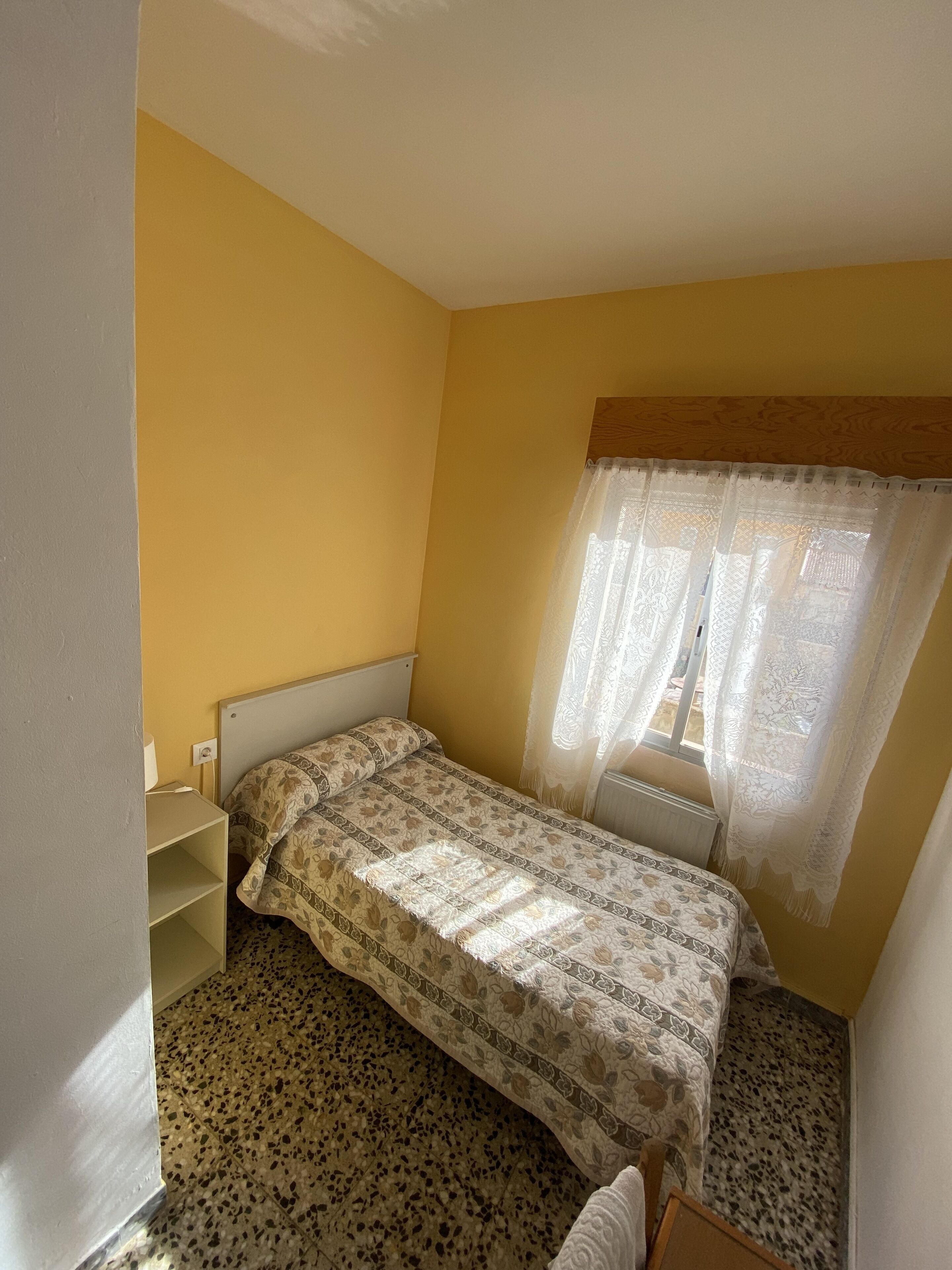 A small, single-bed room with a patterned bedspread, a radiator, and a window with sheer curtains.